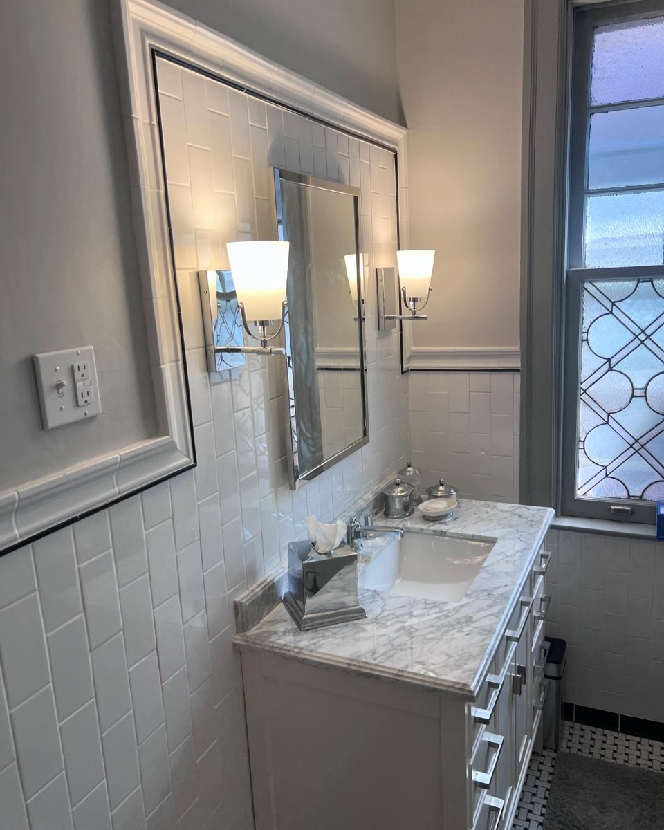 Bright white bathroom remodel with marble-top vanity, wall sconces, and leaded-glass window